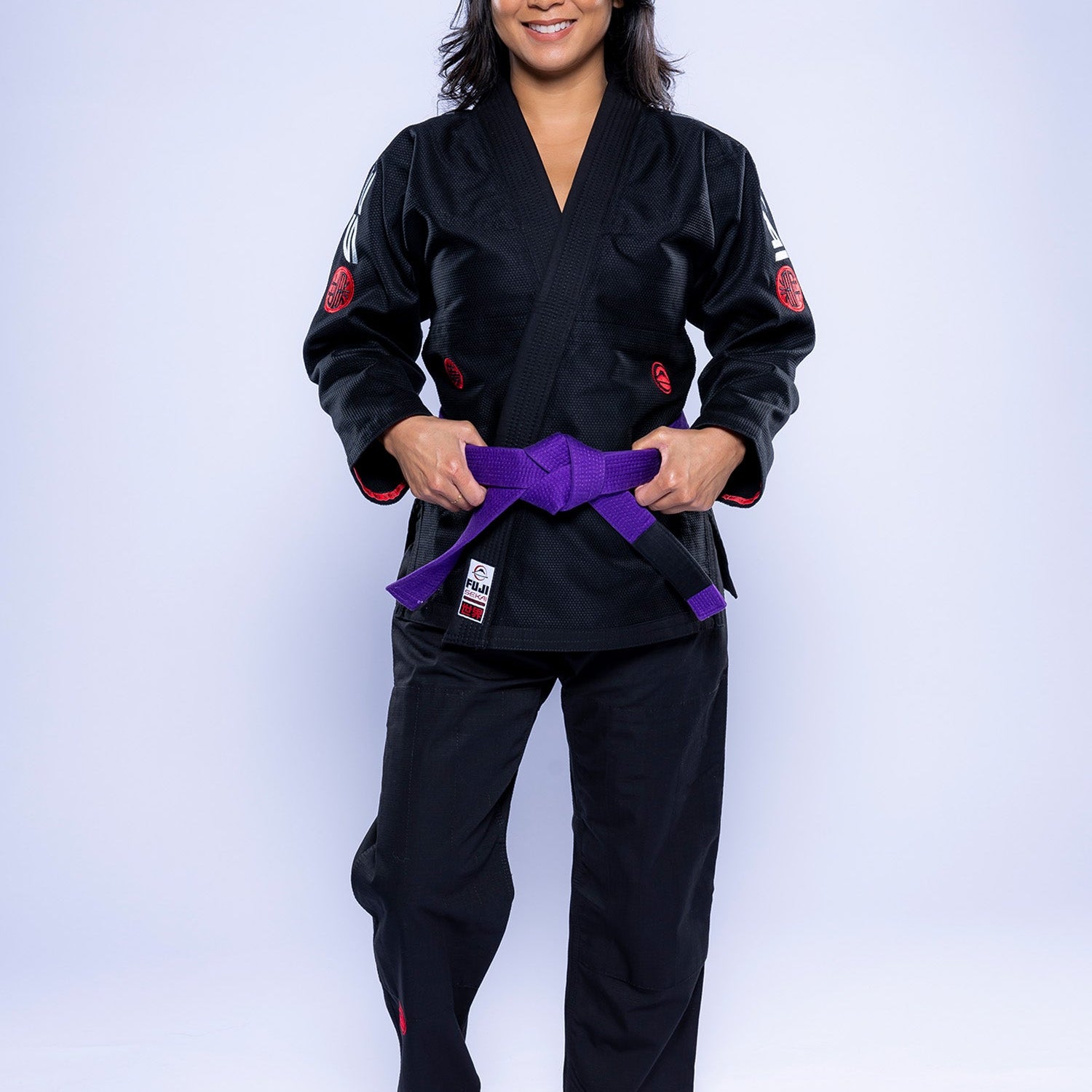 A woman stands barefoot on a white background, smiling and wearing the Womens SEKAI 3.0 BJJ Gi Black Grey with a purple belt tied around her waist.