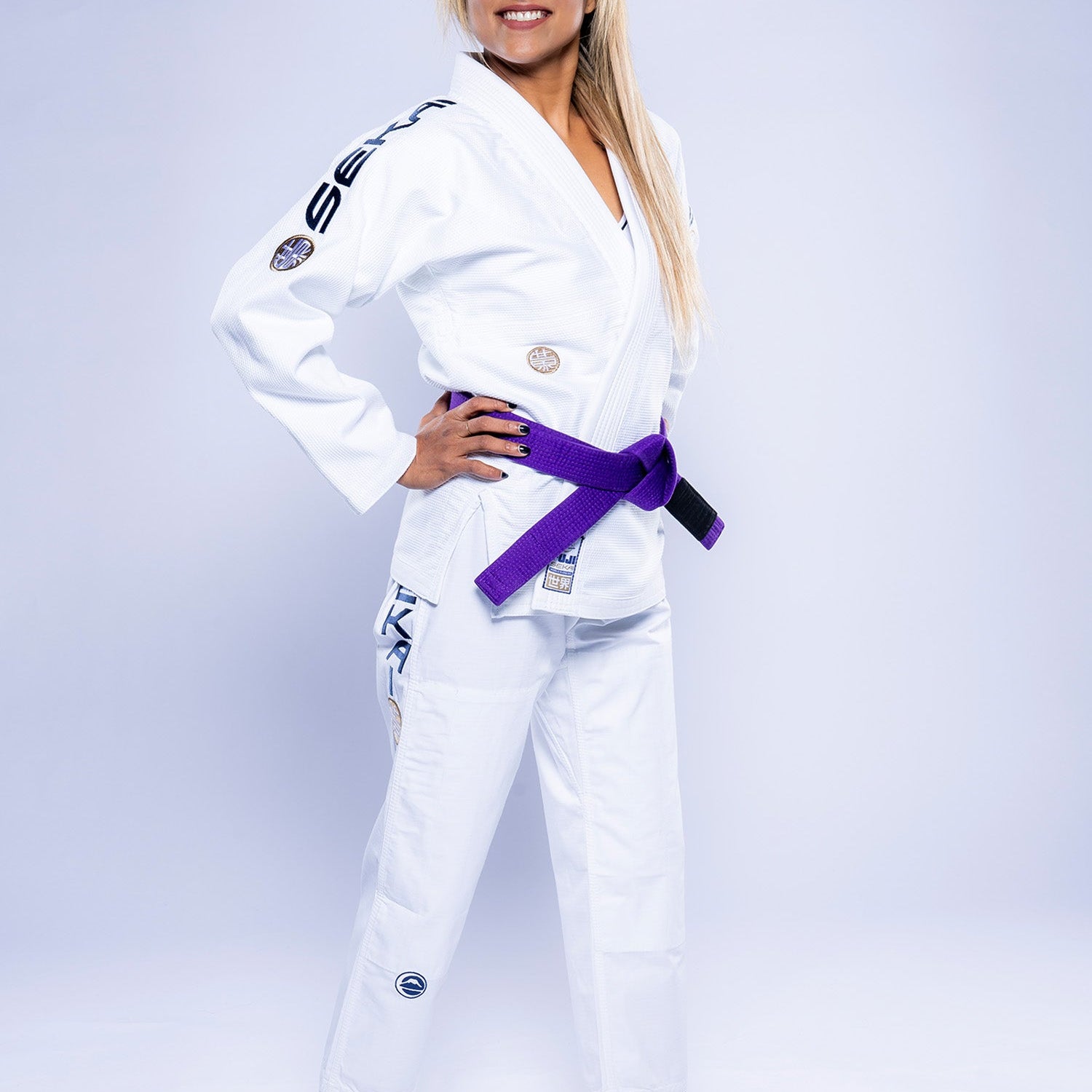 A woman smiles and looks to the side, standing barefoot with hands on her hips in a Womens SEKAI 3.0 BJJ Gi White Navy and a purple belt, against a plain light background.