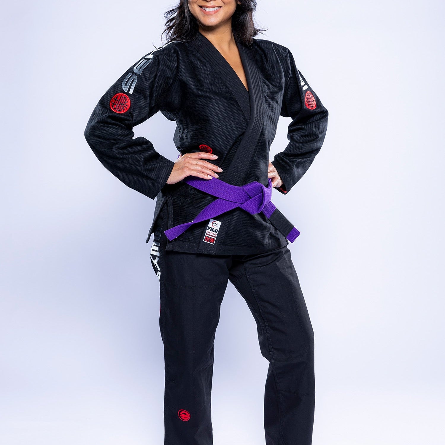 A woman stands barefoot on a white background, smiling with her hands on her hips and wearing the Womens SEKAI 3.0 BJJ Gi Black Grey with a purple belt.