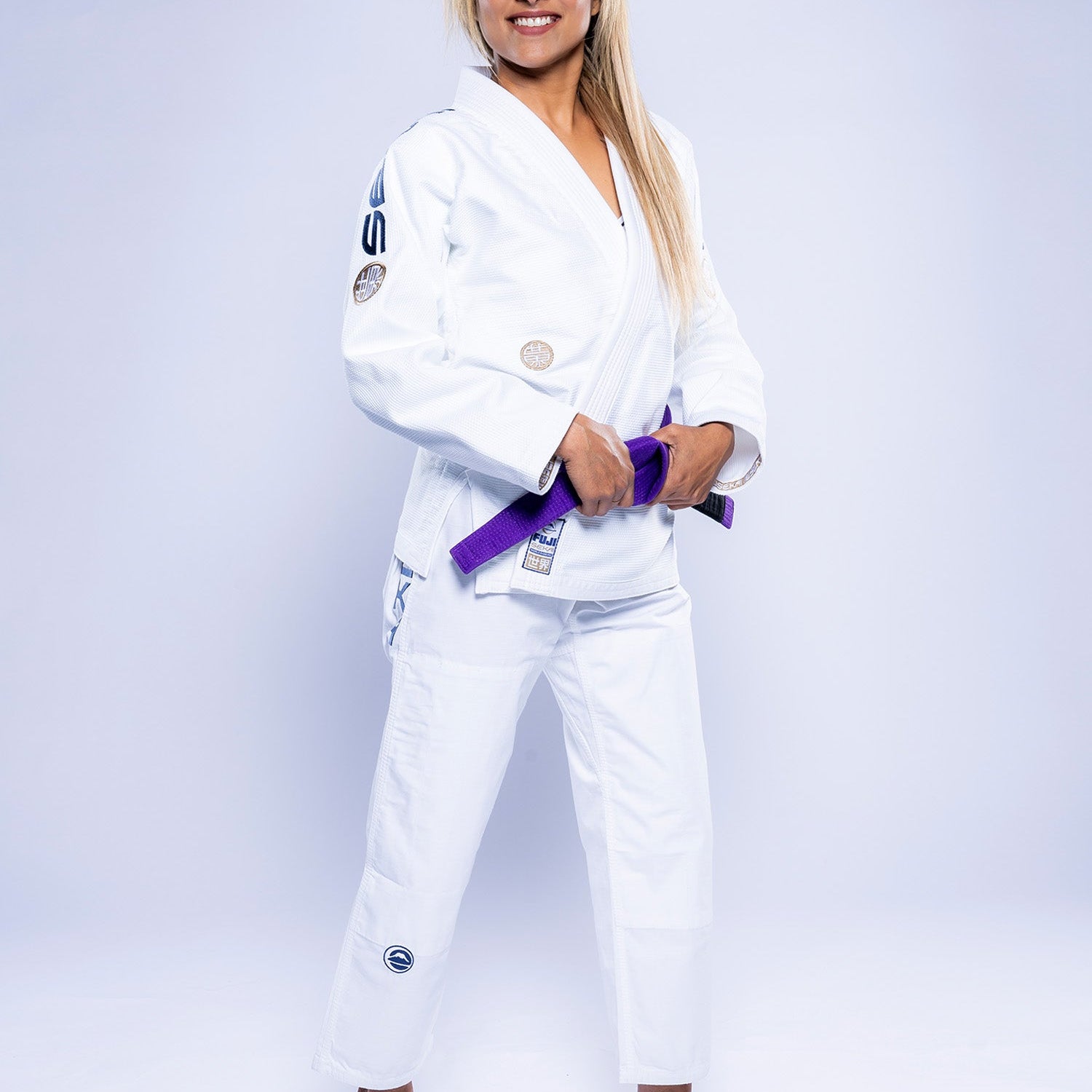 A woman wearing the Womens SEKAI 3.0 BJJ Gi White Navy stands barefoot, smiling and holding a purple belt in front of her against a plain light background.