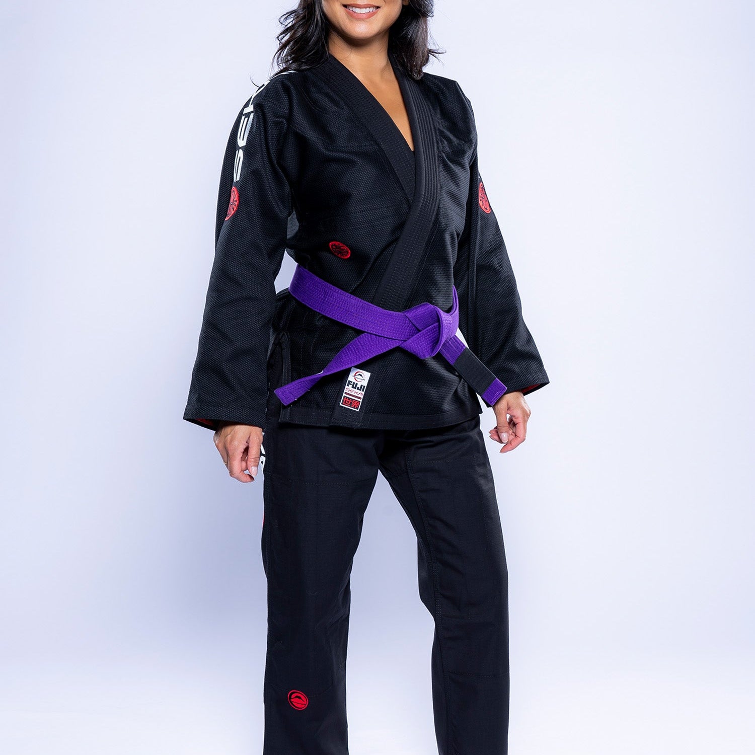A woman stands barefoot in a Womens SEKAI 3.0 BJJ Gi Black Grey with a purple belt, smiling at the camera against a plain white background.
