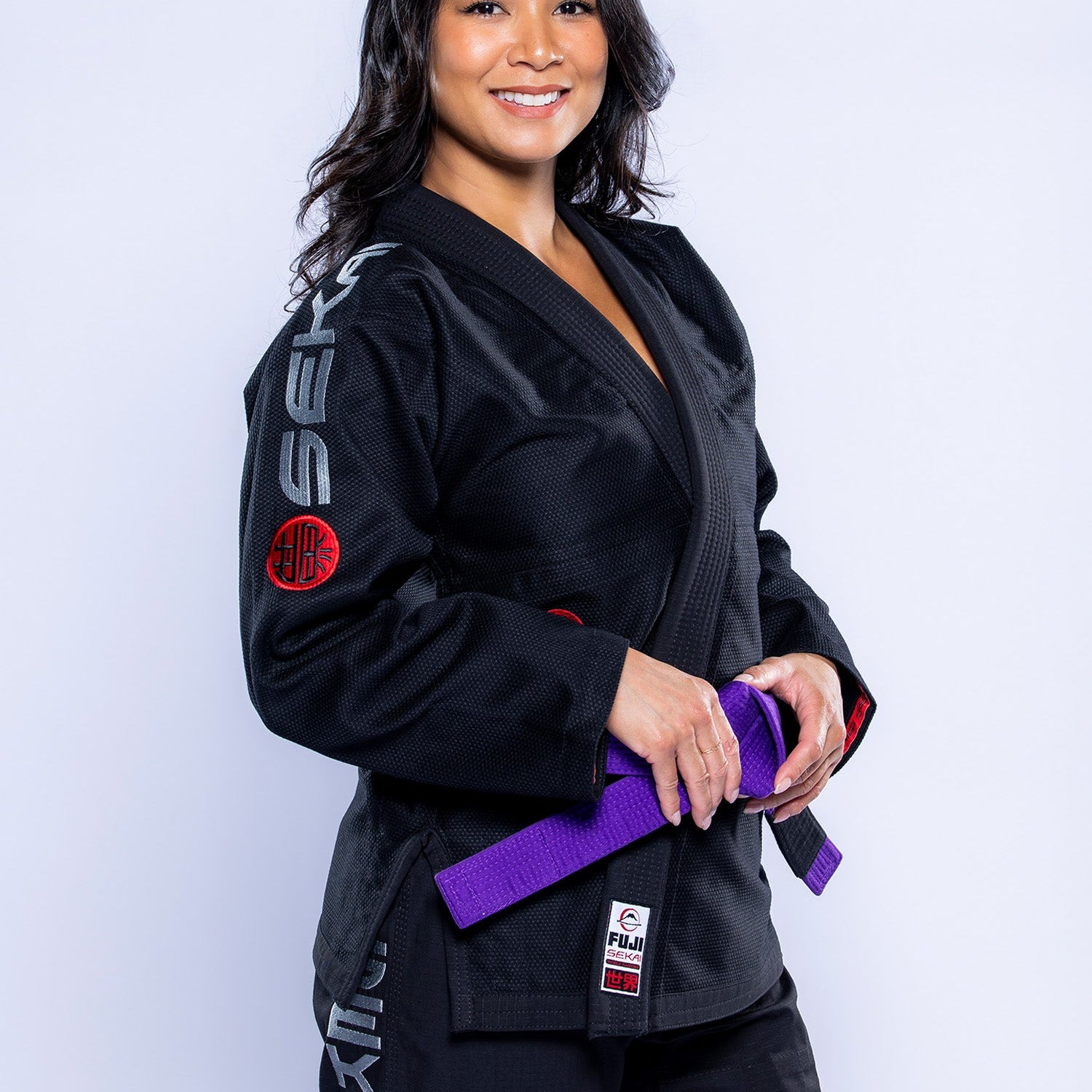 A woman in the Womens SEKAI 3.0 BJJ Gi Black Grey with patches and a purple belt smiles against a plain white background.