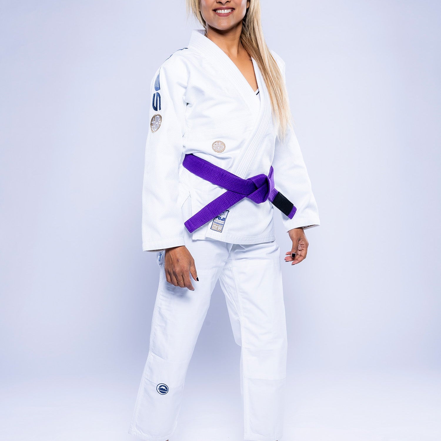 A woman with long loose hair smiles confidently at the camera, barefoot on a light background, wearing the white Women's SEKAI 3.0 BJJ Gi in Navy with a purple belt.