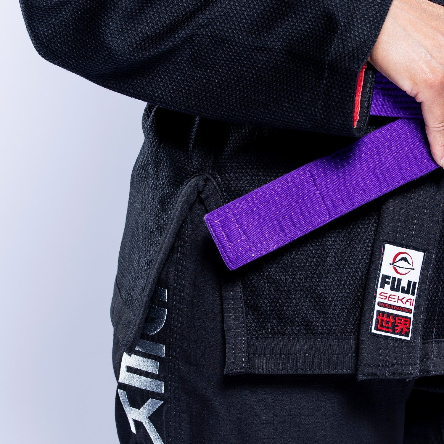 A woman wearing the Womens SEKAI 3.0 BJJ Gi Black Grey with white lettering on the pants and a purple belt adjusts her belt. The Fuji brand patch is visible on the gi.