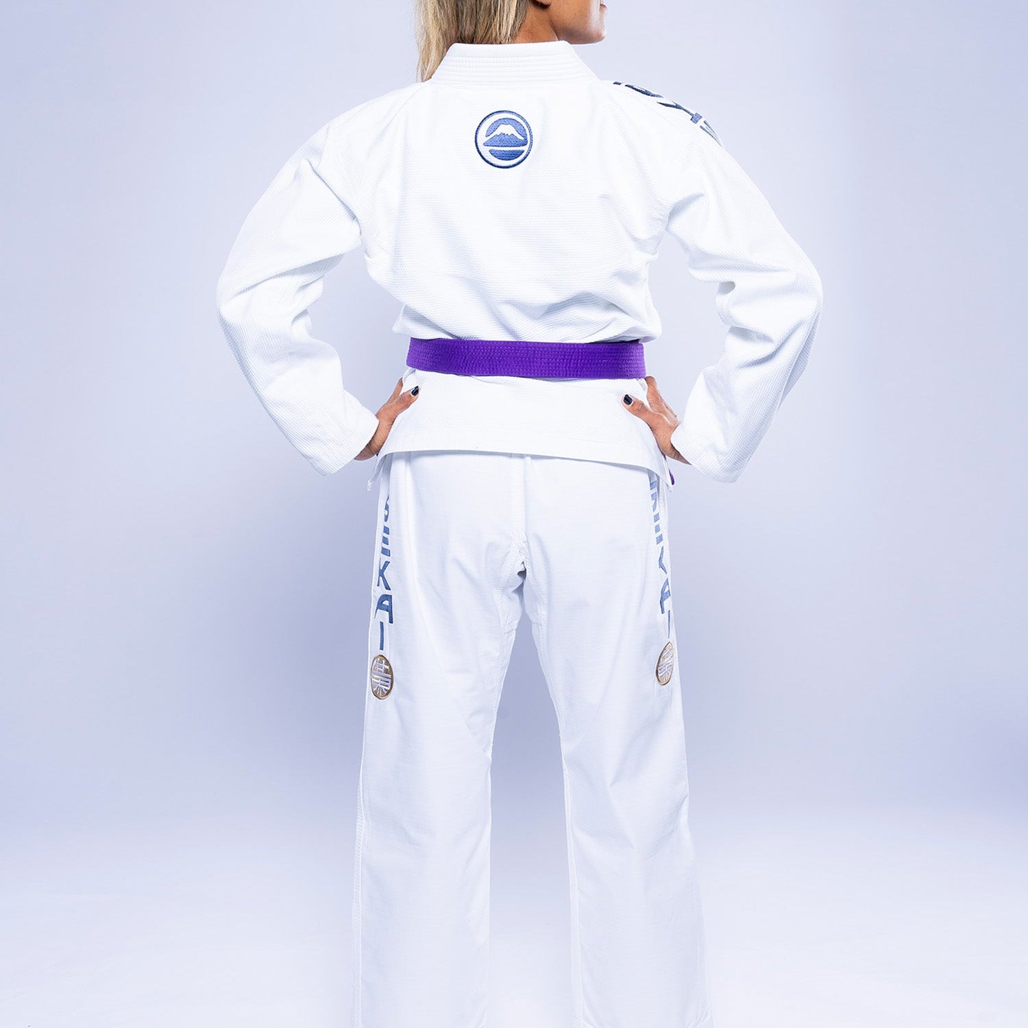 A person with long blonde hair faces away, wearing the Womens SEKAI 3.0 BJJ Gi White Navy with a purple belt. The gi features blue and gray patches and logos on the back and legs, set against a plain light background.