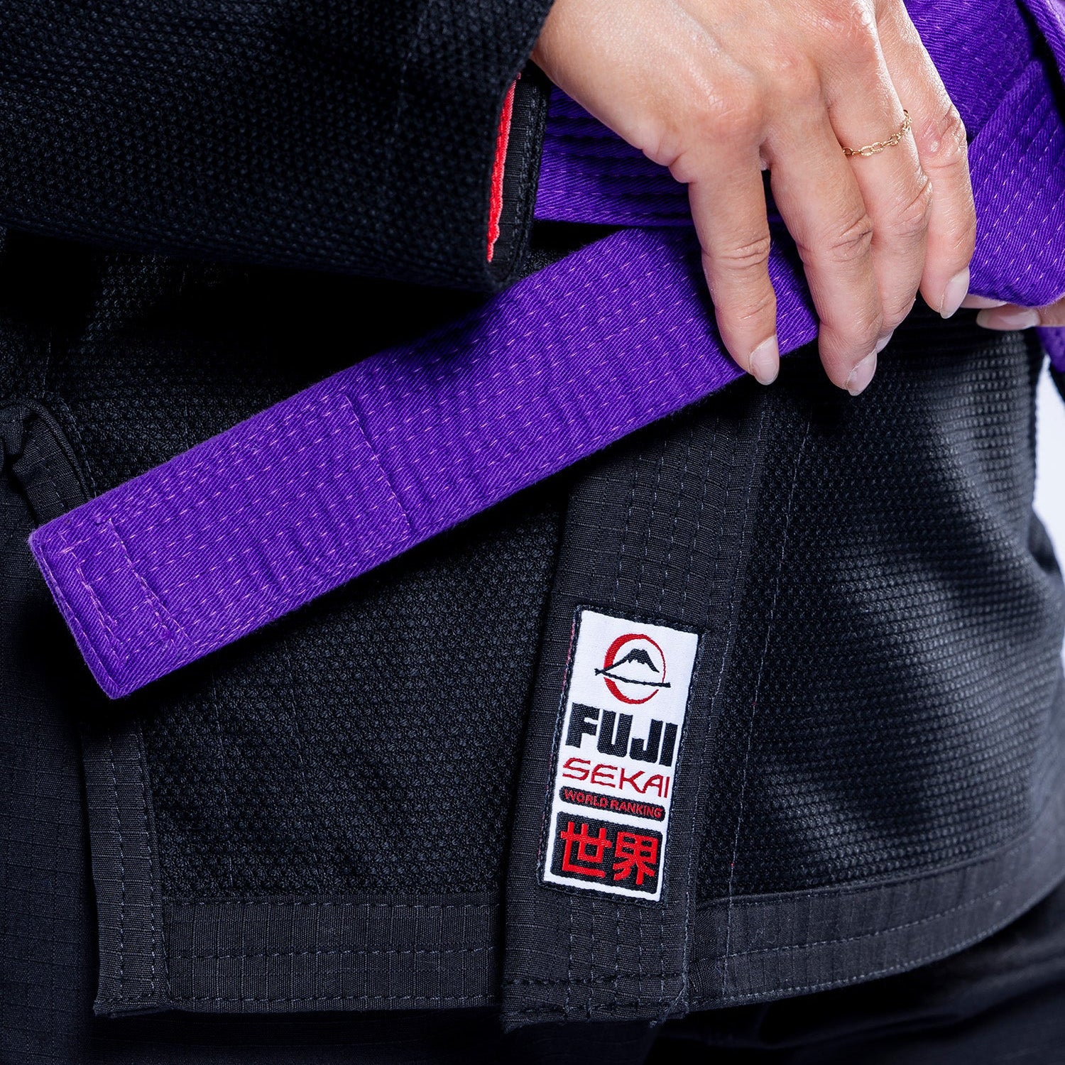 Close-up of a person’s hands tying a purple belt around the waist of the Womens SEKAI 3.0 BJJ Gi Black Grey, featuring a Fuji Seki patch with Japanese characters.