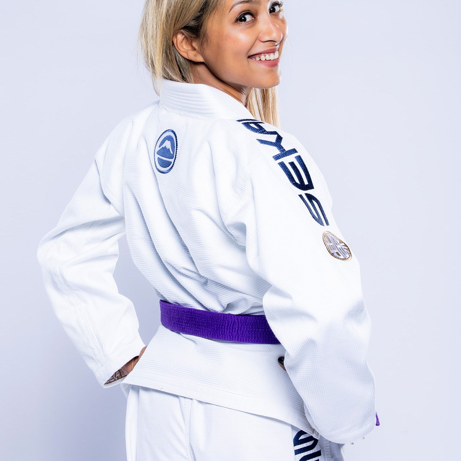 A woman with long blonde hair smiles over her shoulder, wearing the Womens SEKAI 3.0 BJJ Gi White Navy with a purple belt and patches on the sleeves and back, standing against a light background.