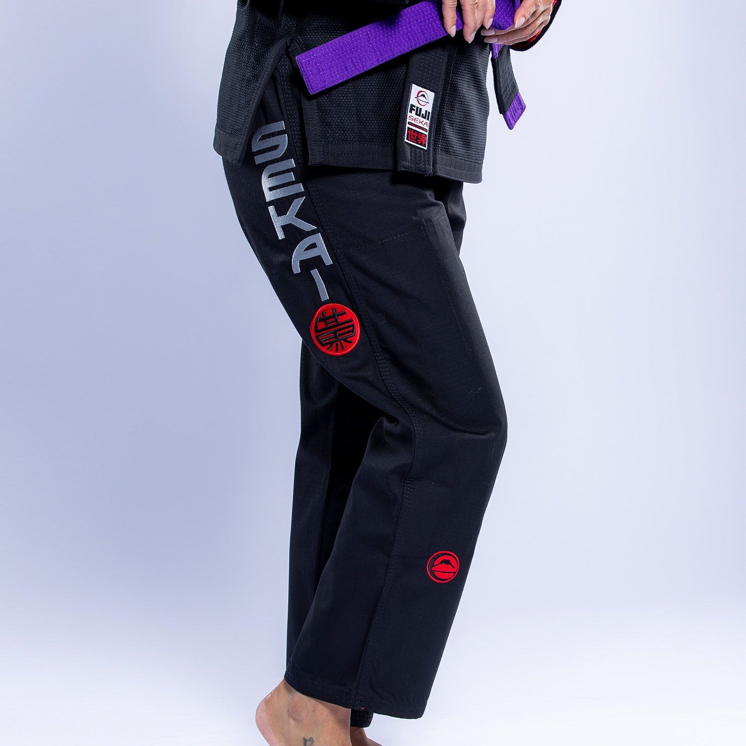A person stands barefoot wearing the Womens SEKAI 3.0 BJJ Gi Black Grey with red patches and "SEKAI" printed on the pants, a purple belt around their waist, and a small tattoo on their left foot.