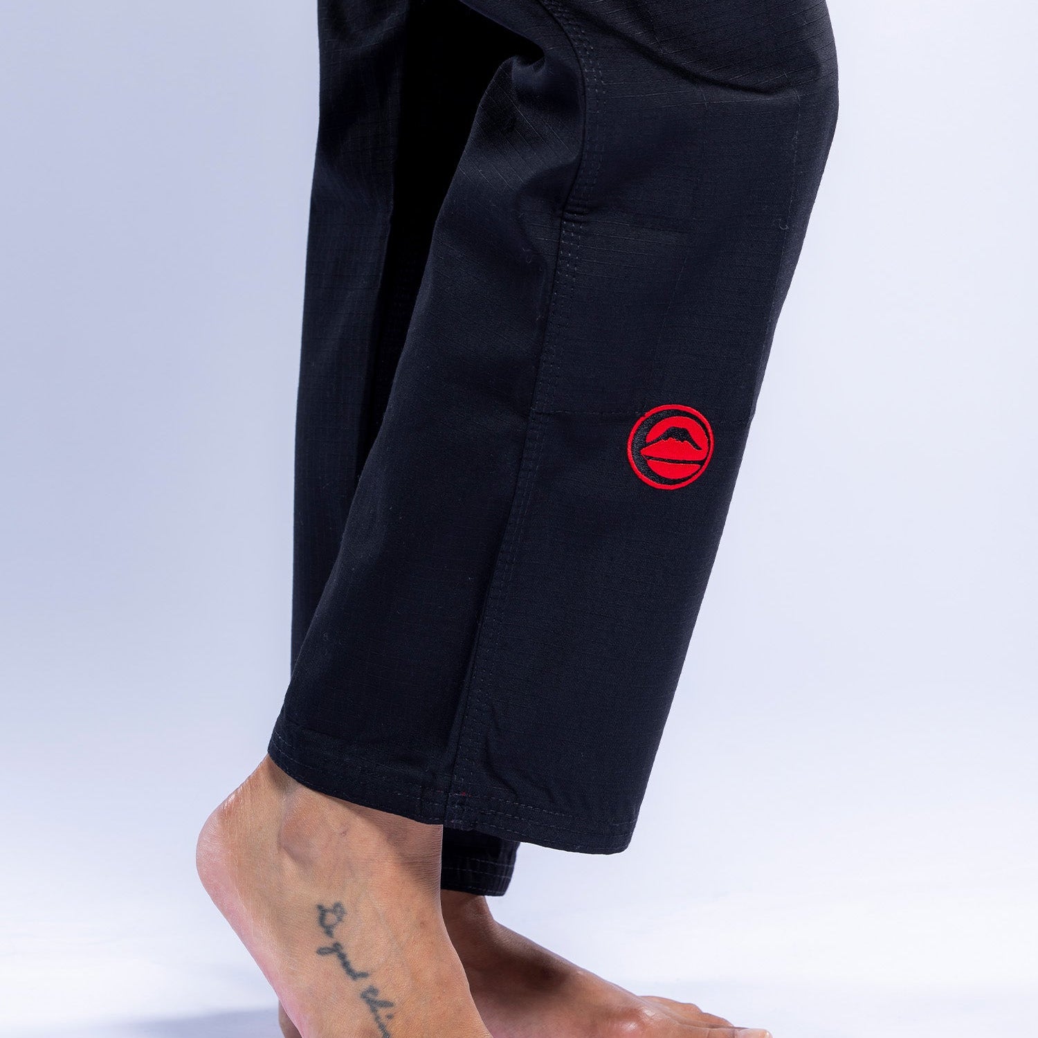 A person stands barefoot on tiptoes in the Womens SEKAI 3.0 BJJ Gi Black Grey, featuring red circular embroidered patches. A star tattoo is visible on the outer left foot against a plain white background.