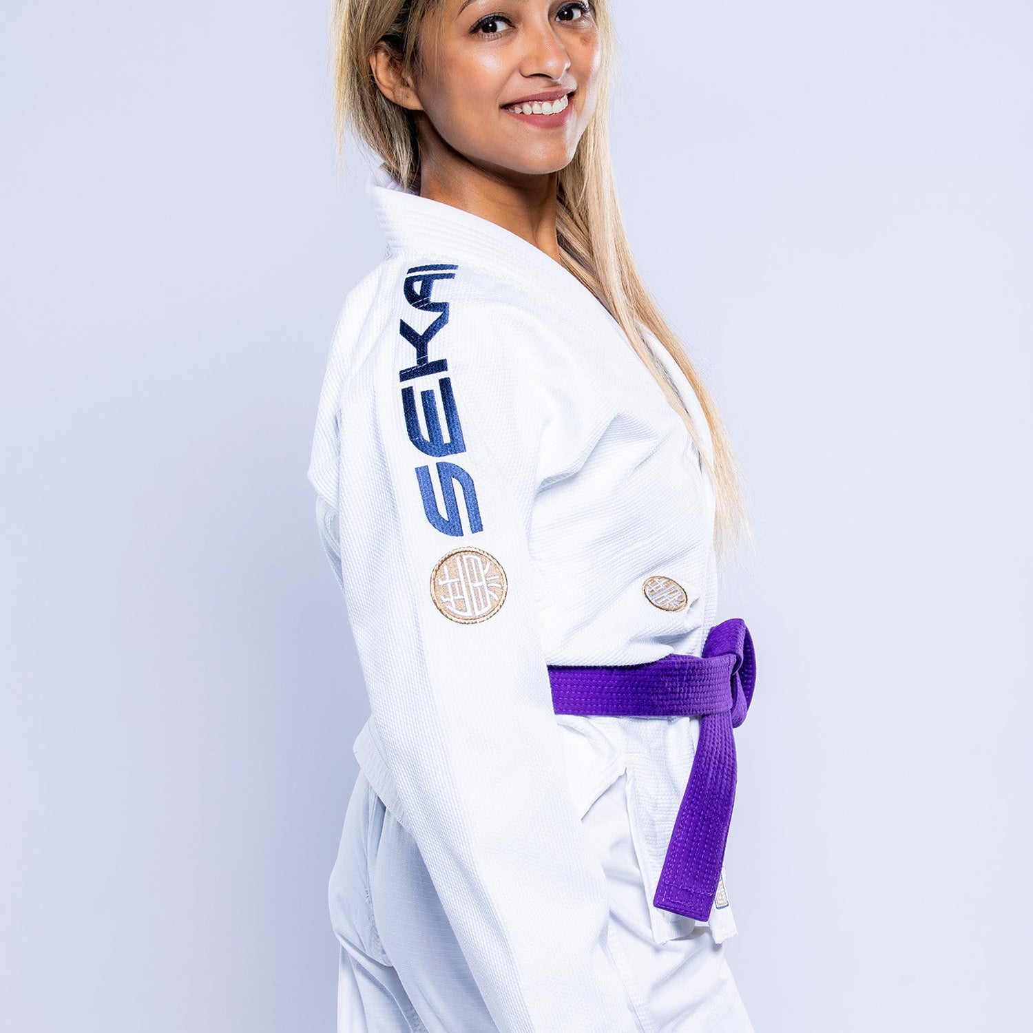 A woman with long blonde hair smiles while wearing the Womens SEKAI 3.0 BJJ Gi White Navy with a purple belt. SEKA is written on the shoulder, and patches are on the chest and arm. The background is plain light gray.
