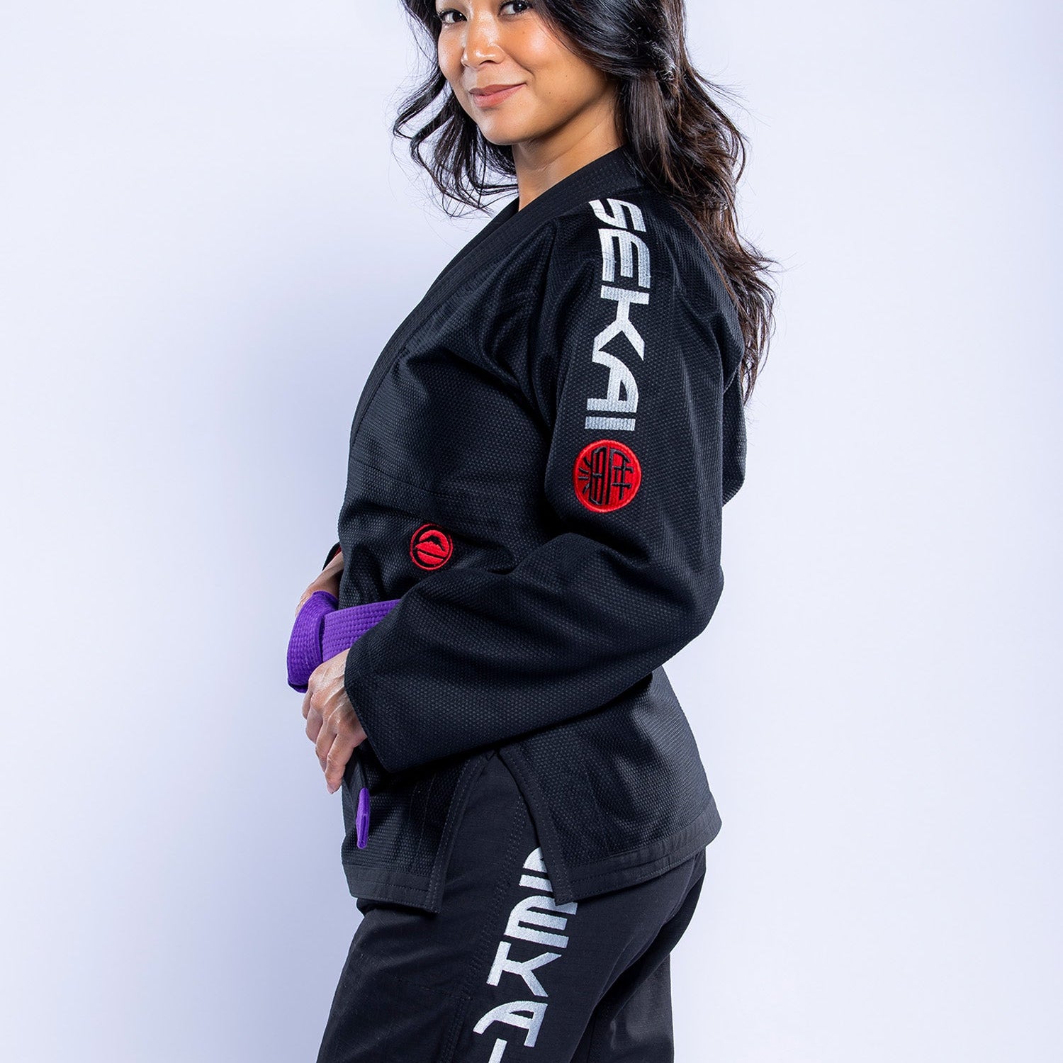 A woman with long dark hair wears the Womens SEKAI 3.0 BJJ Gi Black Grey, featuring SEKA on the sleeves and pants. She has a purple belt and smiles slightly at the camera against a light background.