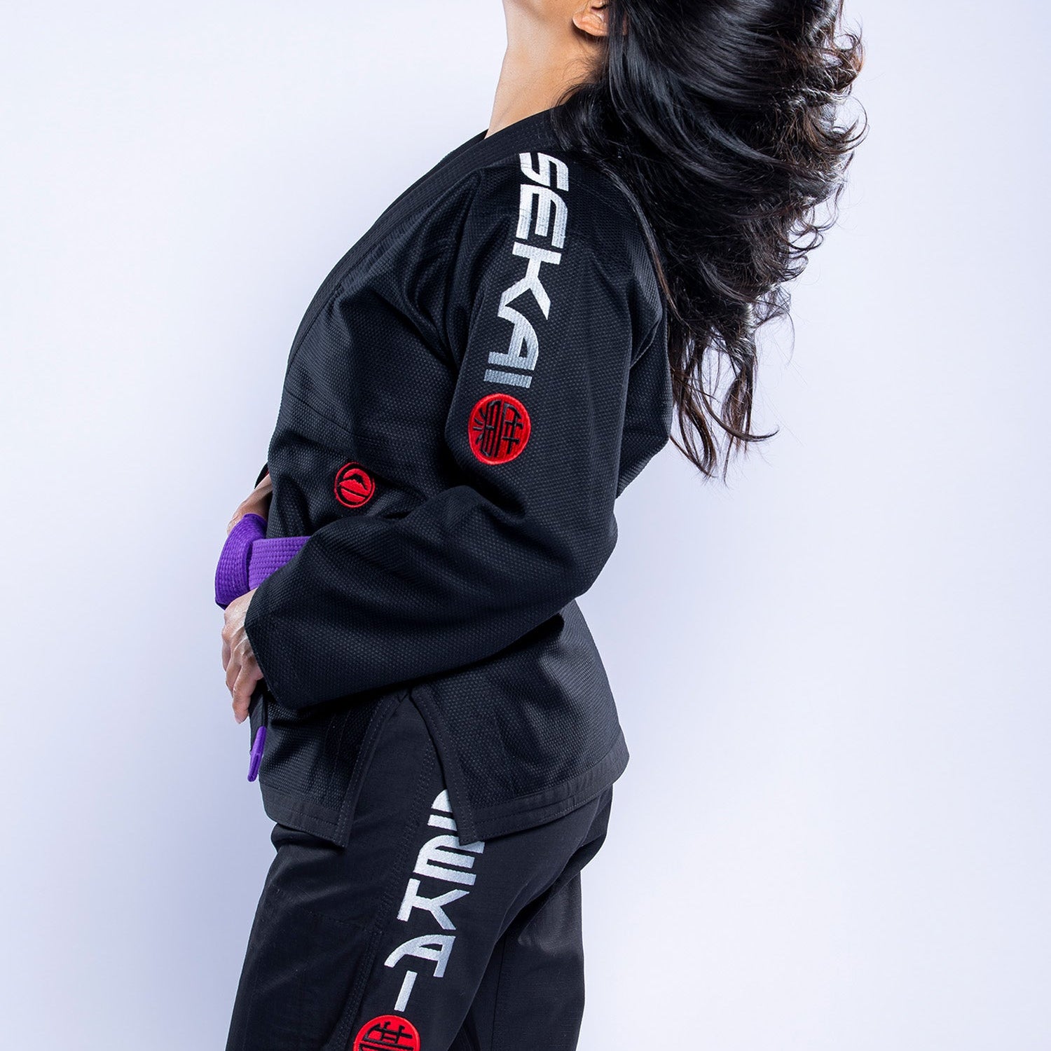 A woman in the Womens SEKAI 3.0 BJJ Gi Black Grey stands in profile, holding a purple belt and tossing her hair back, with "SEKAI" text and red patches visible on the gi.