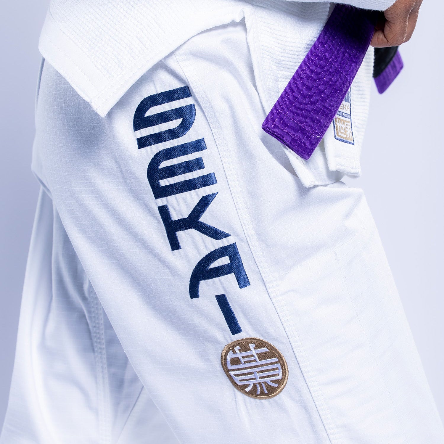 Close-up of a person wearing the Womens SEKAI 3.0 BJJ Gi White Navy with black and gold embroidery on the pants and a purple belt at the waist.