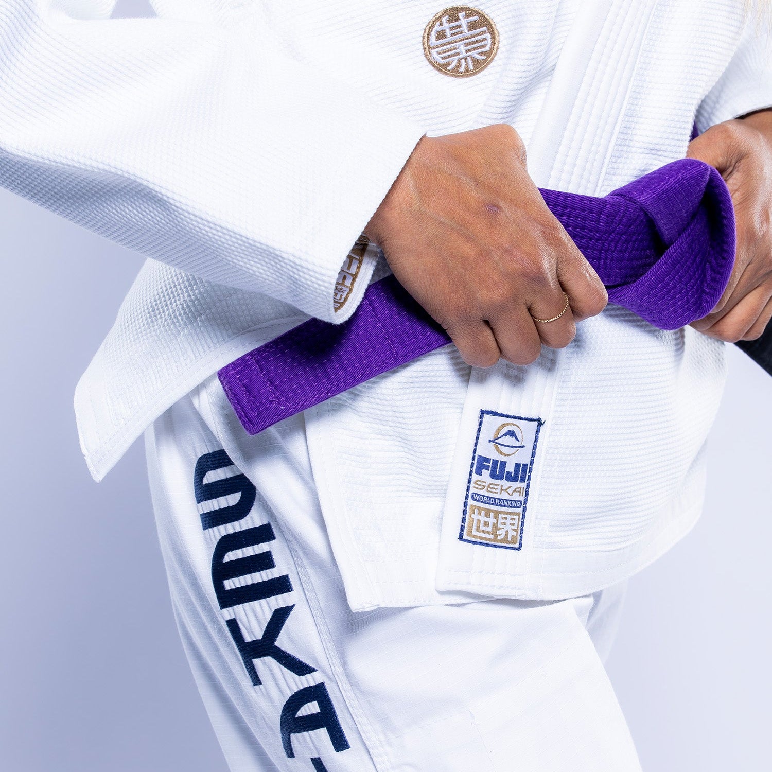 A person wears the Womens SEKAI 3.0 BJJ Gi White Navy with patches and ties a purple belt around their waist. The gi displays Japanese characters and the FUJI SEKAI logo on the jacket and pants.