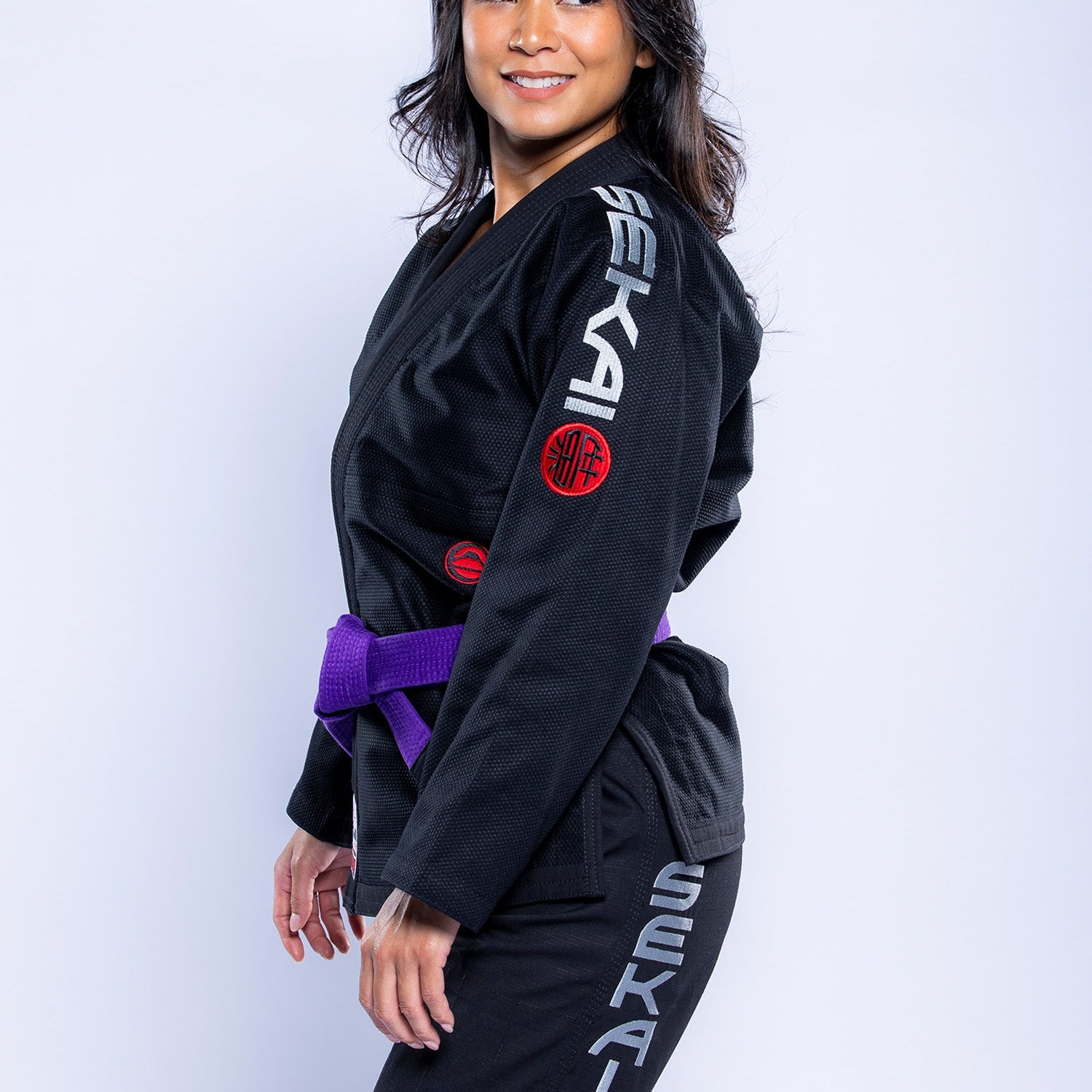 A woman in a black and grey Womens SEKAI 3.0 BJJ Gi with red patches and a purple belt stands sideways, smiling against a plain light background.