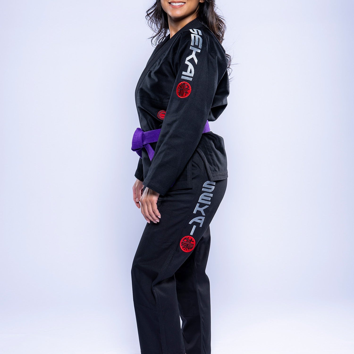 A woman wears the Womens SEKAI 3.0 BJJ Gi Black Grey, featuring SEKAI on the sleeve and pant leg, a purple belt, and stands barefoot smiling against a white background.