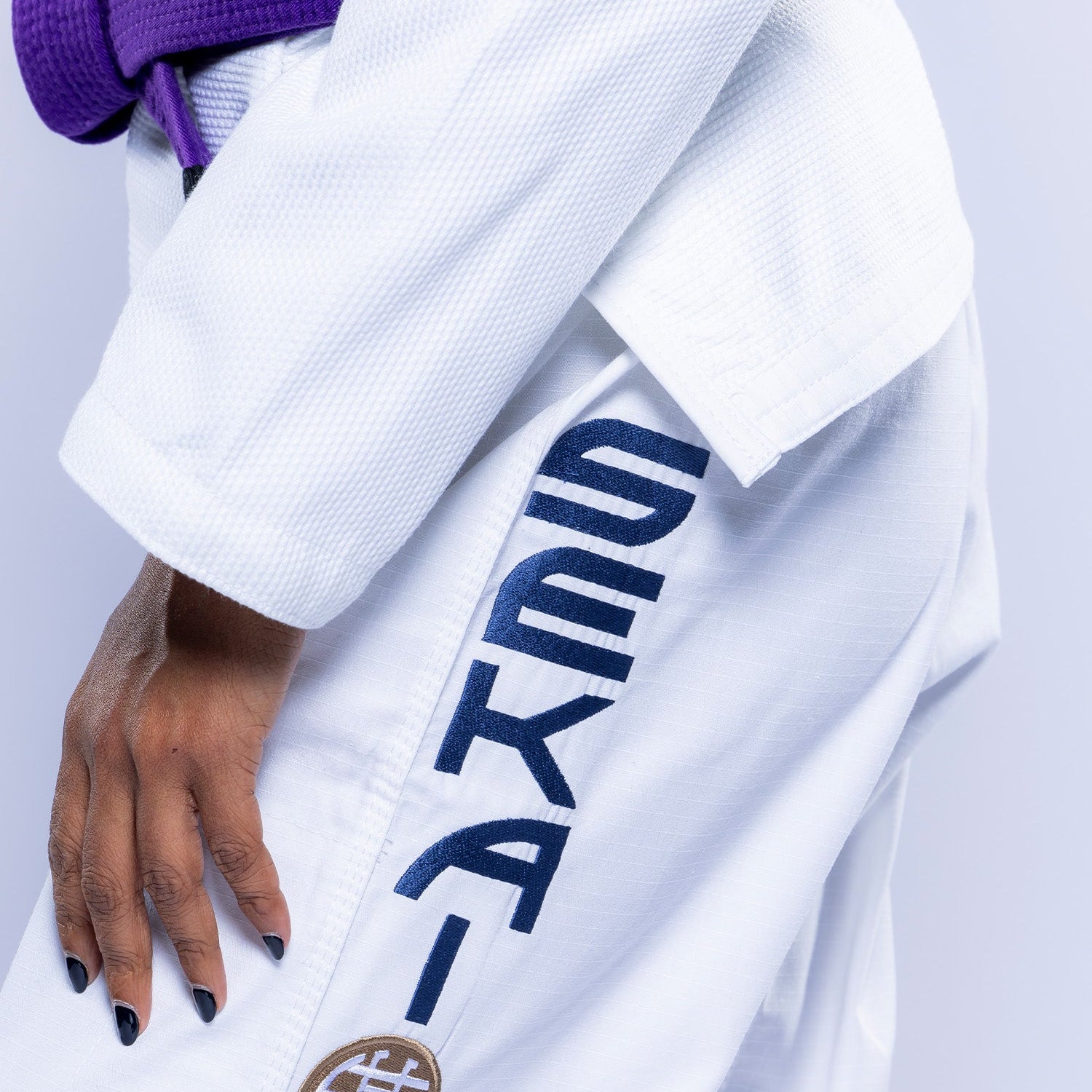 A person wears the Womens SEKAI 3.0 BJJ Gi White Navy, featuring "SEKAI" in bold and a circular patch with Japanese characters on the pants, paired with a purple belt.