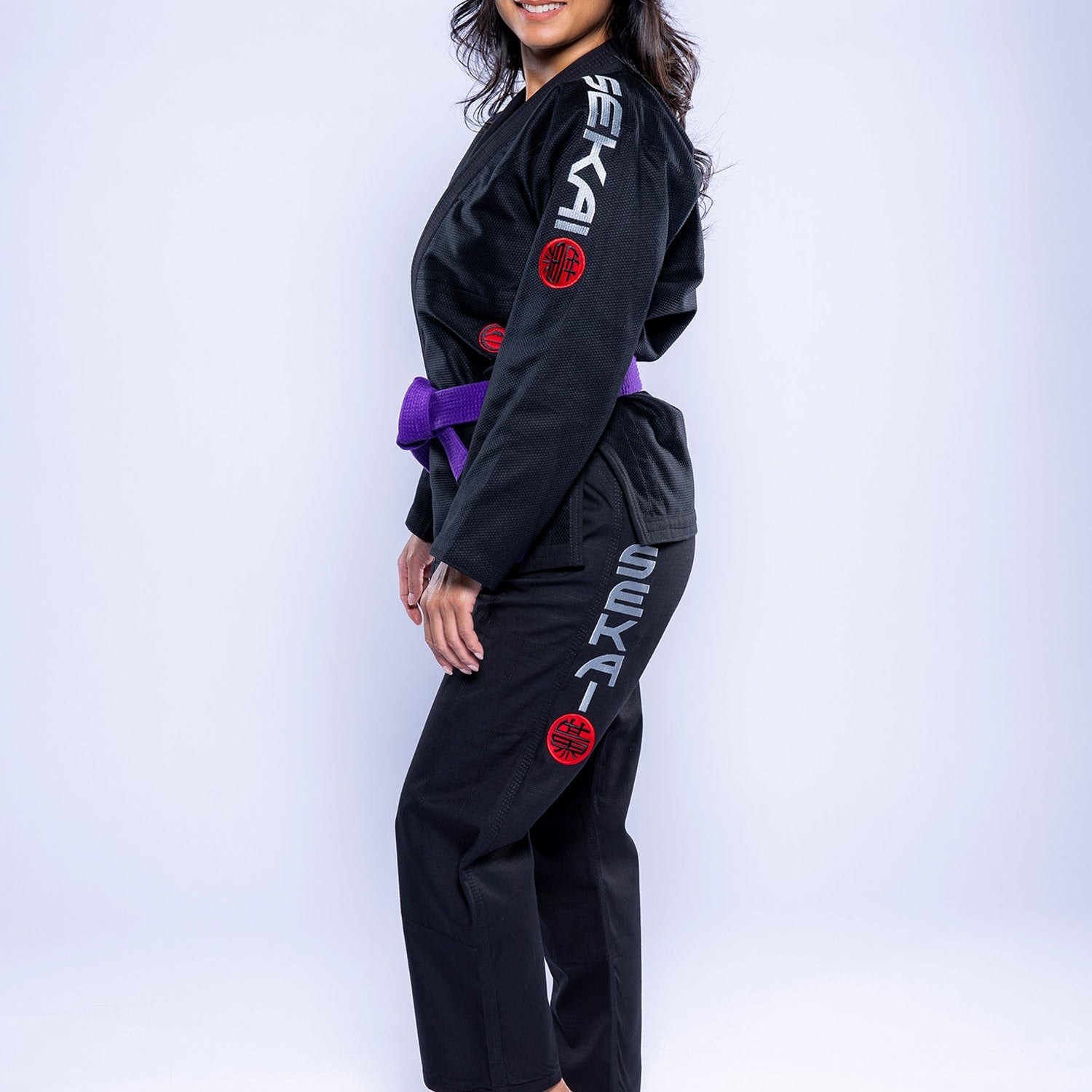 A woman wearing the Womens SEKAI 3.0 BJJ Gi Black Grey with a purple belt stands barefoot, smiling and posing against a plain white background. The gi features gray SEKA lettering and red circular patches on the sleeve and pants.