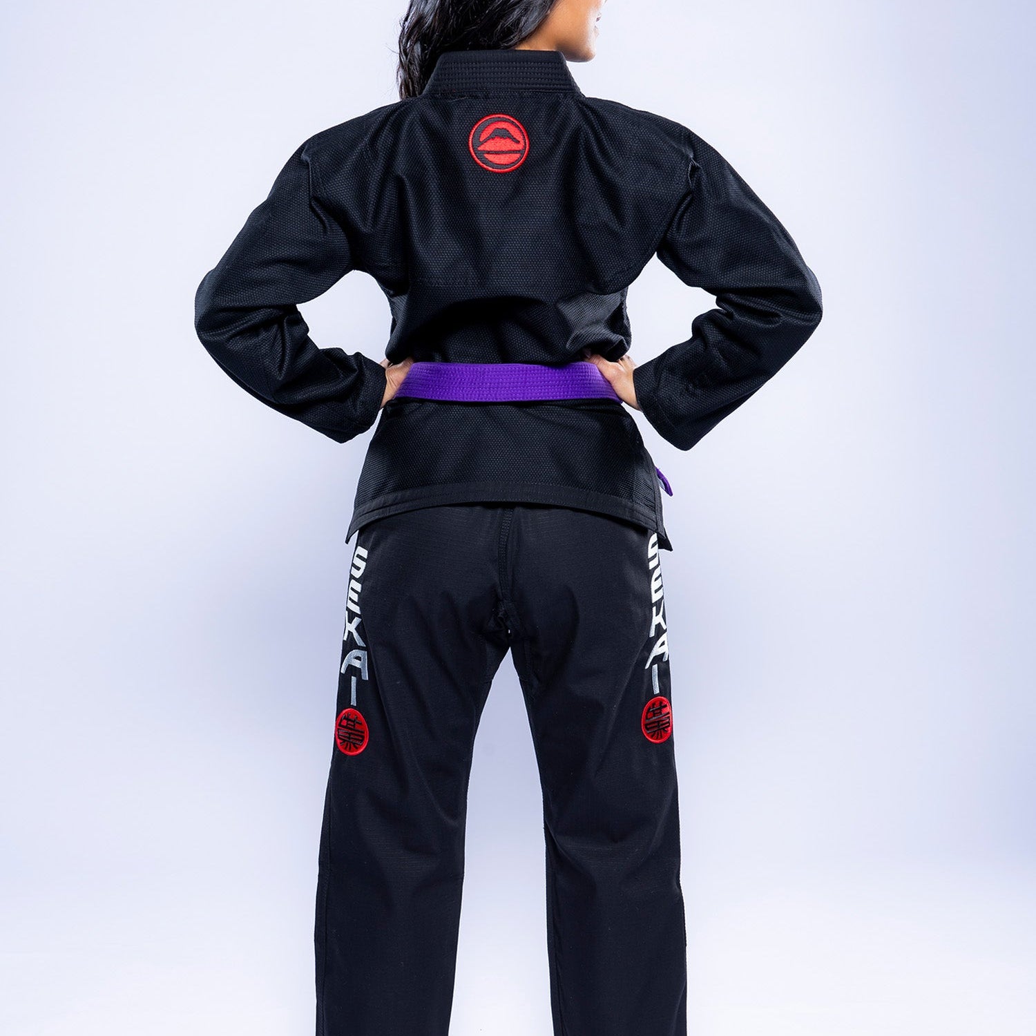 A person with long dark hair stands barefoot, facing away, wearing the Womens SEKAI 3.0 BJJ Gi Black Grey adorned with a purple belt. Red and white patches are seen on the back and legs of the uniform.
