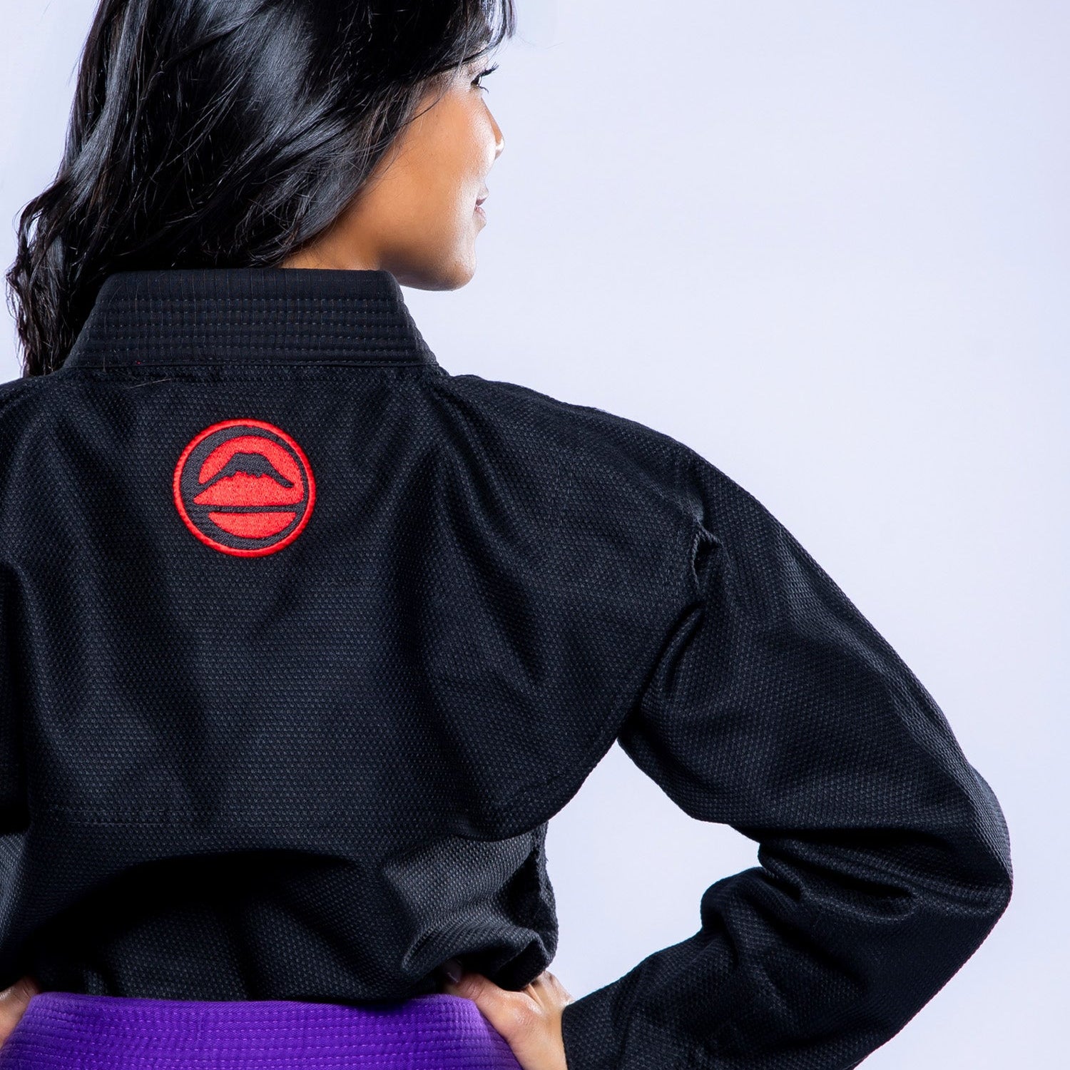 A woman with long dark hair stands facing away from the camera, hands on hips, wearing the Womens SEKAI 3.0 BJJ Gi Black Grey and a purple belt, against a light background.