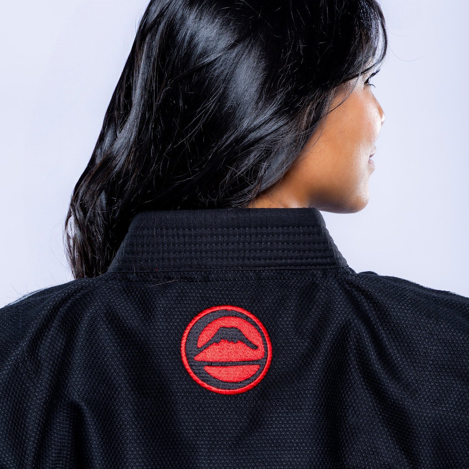 A woman with long dark hair is shown from behind, wearing the Womens SEKAI 3.0 BJJ Gi Black Grey, which features a red circular patch with a mouth and mustache on the back of the collar.