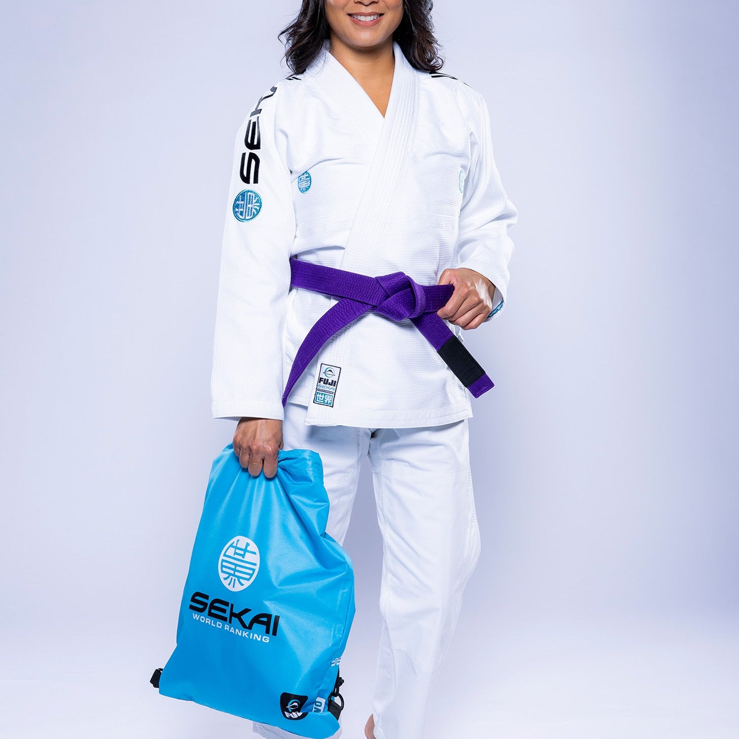 A woman in a white and teal Womens SEKAI 3.0 BJJ Gi with a purple belt stands barefoot, smiling, and holds a blue Sekai sports bag against a plain light background.
