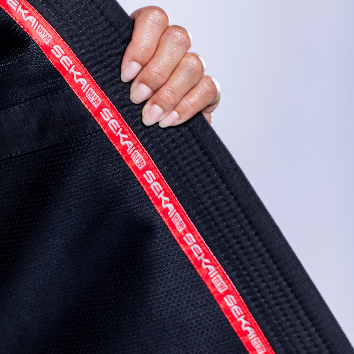 A hand holds the collar of the Womens SEKAI 3.0 BJJ Gi Black Grey, featuring black fabric with red trim and "SEKAI" repeatedly printed in white. The background is a soft, light gray.