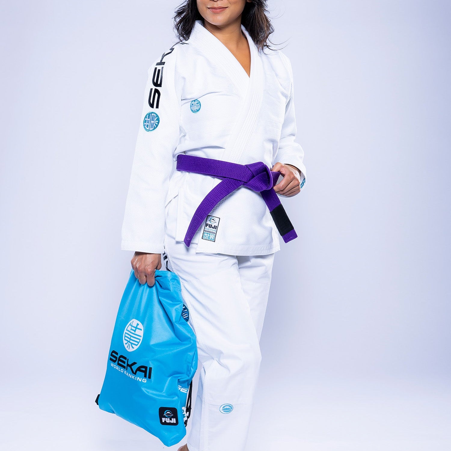 A woman wearing the Womens SEKAI 3.0 BJJ Gi White Teal with a purple belt stands barefoot on a light background, confidently smiling at the camera while holding a light blue bag.