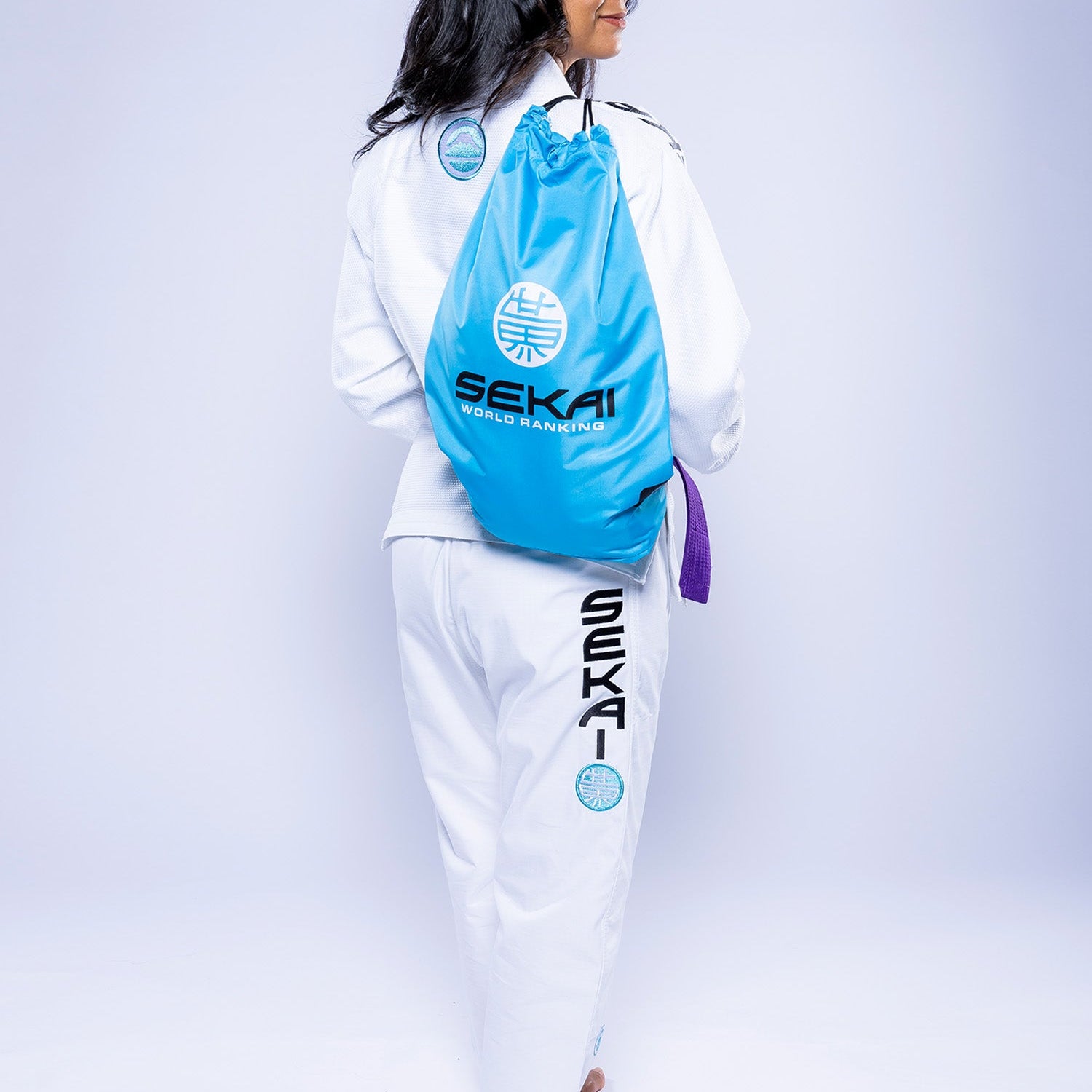A woman in a white Womens SEKAI 3.0 BJJ Gi with teal accents and a purple belt stands barefoot, carrying a bright blue “SEKAI” logo drawstring bag. The background is plain and light.
