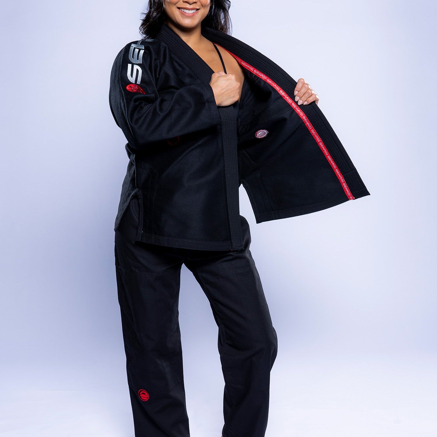 A woman smiles at the camera, barefoot in the Womens SEKAI 3.0 BJJ Gi Black Grey, holding open her jacket to display the inner lining against a plain light gray background.