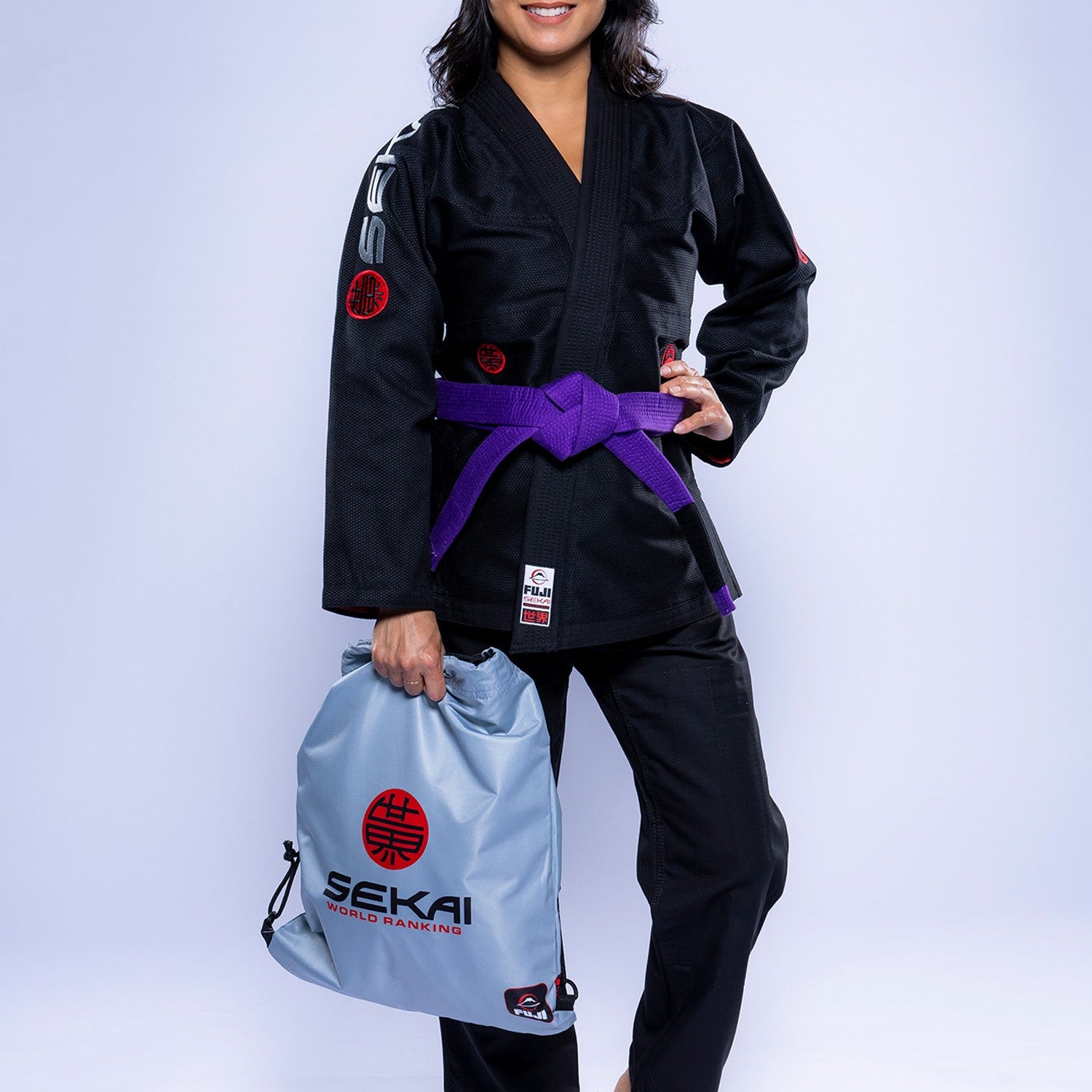 A woman in the Womens SEKAI 3.0 BJJ Gi Black Grey with a purple belt smiles barefoot, holding a light gray drawstring bag featuring red and black SEKAI branding against a plain white background.