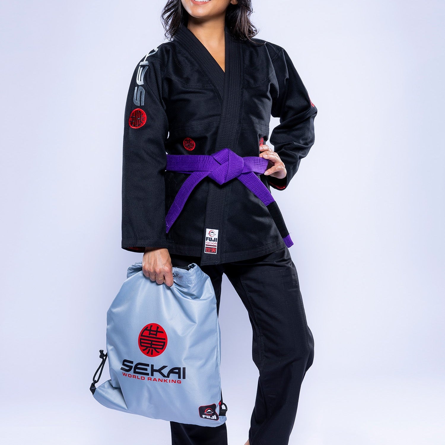 A woman wearing the Womens SEKAI 3.0 BJJ Gi Black Grey and a purple belt stands barefoot, smiling and holding a gray gym bag with SEKAI World Hanegin branding.