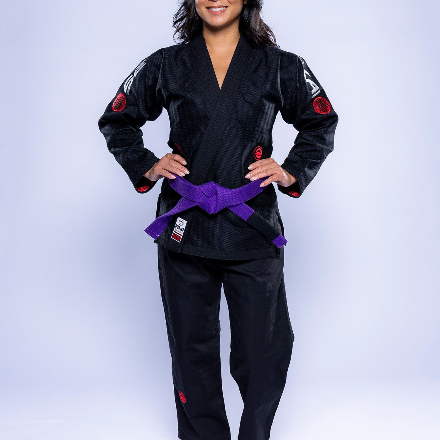 A woman with long dark hair stands barefoot, smiling with hands on hips, wearing the Womens SEKAI 3.0 BJJ Gi in black grey with a purple belt, against a light background.