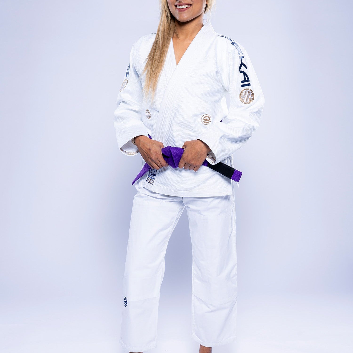 A woman stands barefoot on a white background, smiling and wearing the Womens SEKAI 3.0 BJJ Gi White Navy, with a purple belt tied at her waist.