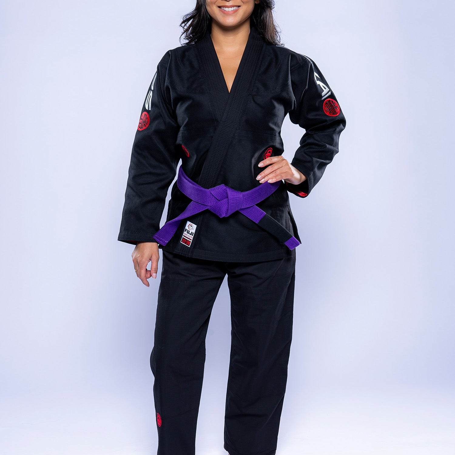 A woman stands barefoot on a white background, smiling and wearing the Womens SEKAI 3.0 BJJ Gi Black Grey with a purple belt tied at her waist, her hands resting at her sides.