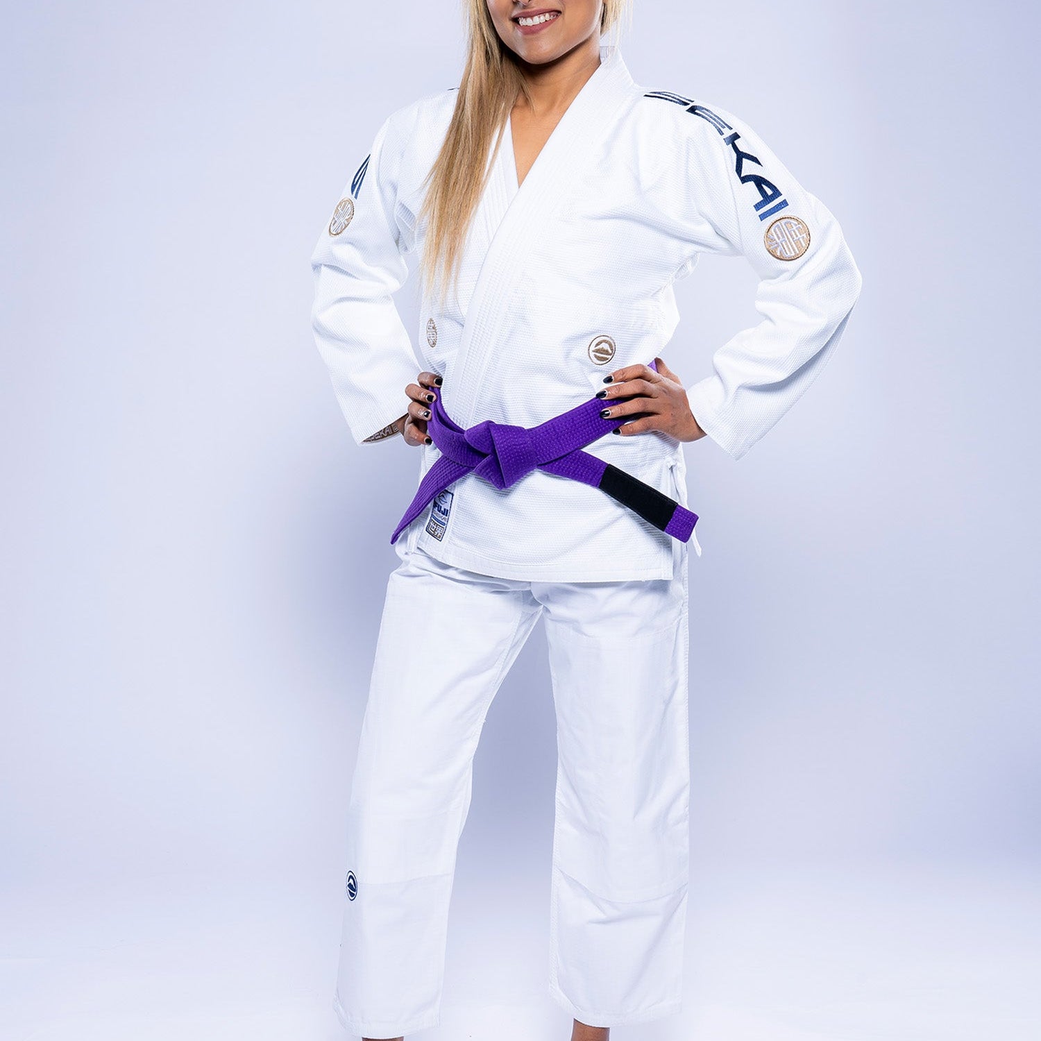 A woman stands confidently, smiling with hands on hips, wearing the Womens SEKAI 3.0 BJJ Gi White Navy and a purple belt against a plain light background.