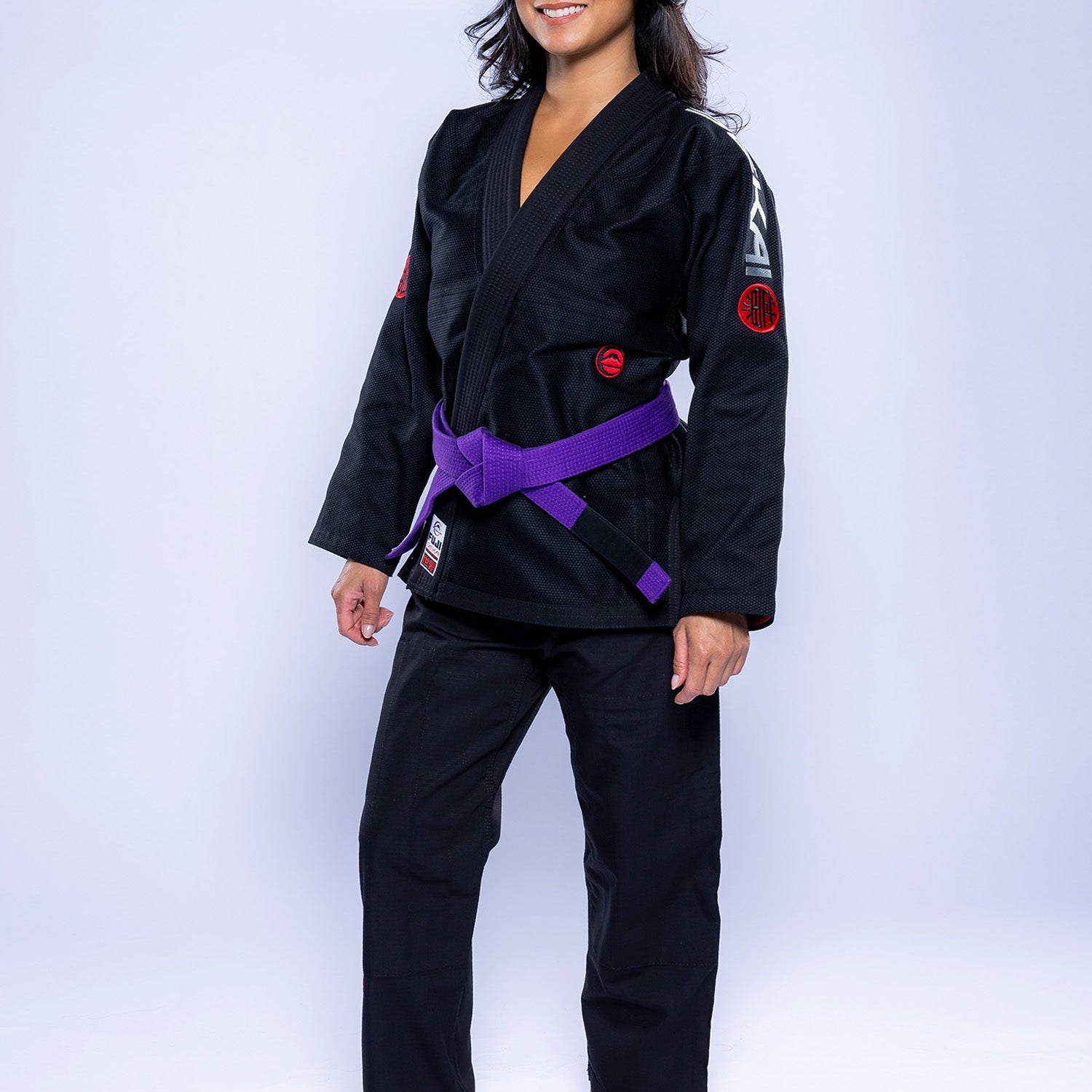 A woman stands barefoot on a light background, smiling and wearing the Womens SEKAI 3.0 BJJ Gi Black Grey with a purple belt tied at her waist.