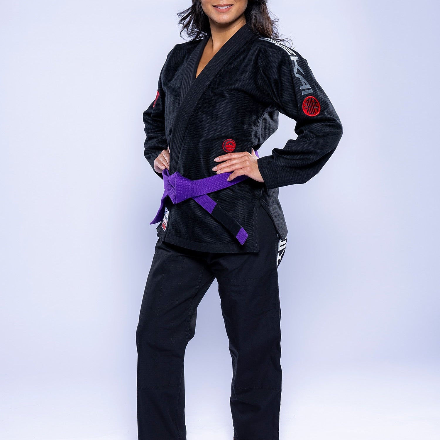 A woman stands barefoot on a white background, wearing the Womens SEKAI 3.0 BJJ Gi Black Grey and a purple belt. She faces forward confidently with hands on hips and a relaxed expression.