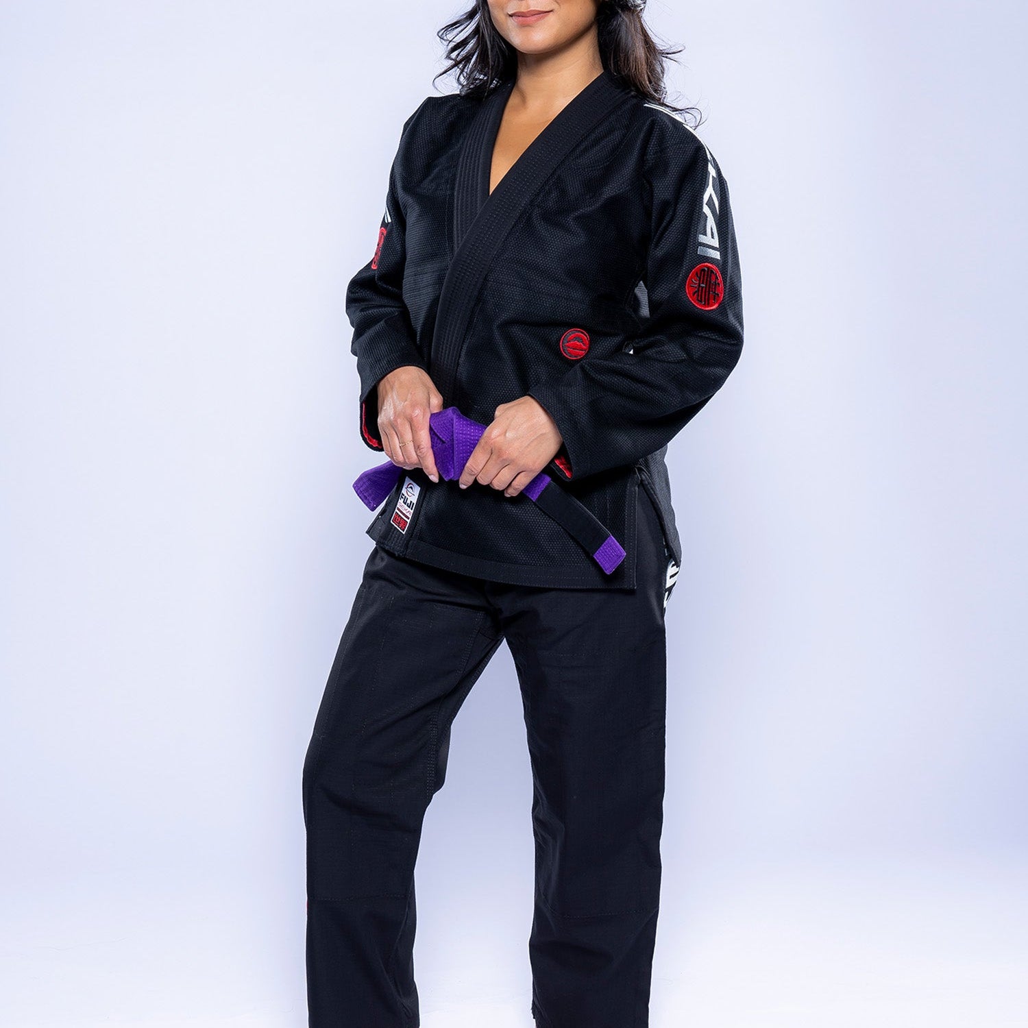 A woman wearing the Womens SEKAI 3.0 BJJ Gi Black Grey holds a purple belt in front of her, barefoot on a white background, looking at the camera with a slight smile.
