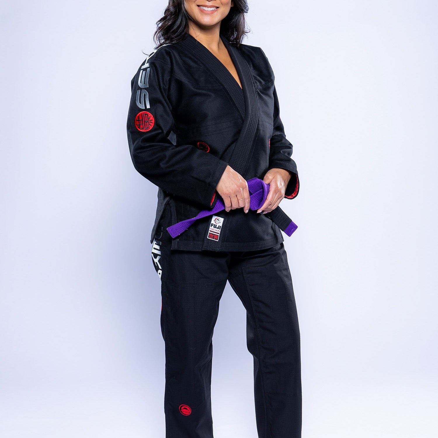 A woman stands barefoot on a white background, smiling and holding a purple belt. She is wearing the Womens SEKAI 3.0 BJJ Gi Black Grey, featuring red and white patches on the sleeves, pants, and chest.