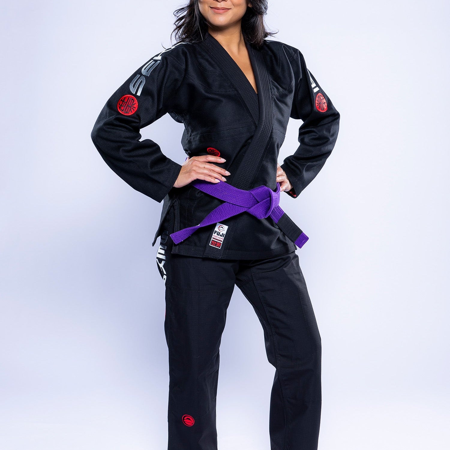 A woman stands barefoot on a white background, wearing the Womens SEKAI 3.0 BJJ Gi Black Grey with a purple belt. She has long dark hair, hands on hips, and looks confidently to the side.