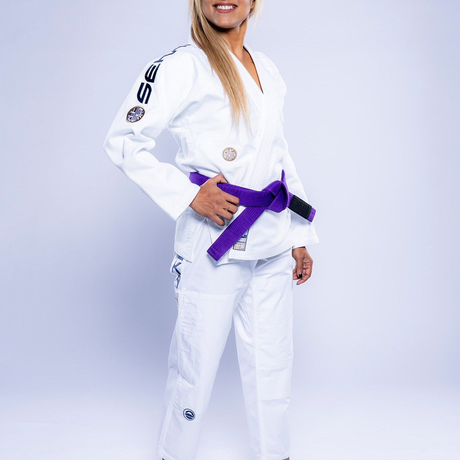 A woman smiles barefoot on a white backdrop, wearing the Womens SEKAI 3.0 BJJ Gi in white/navy with a purple belt tied around her waist.