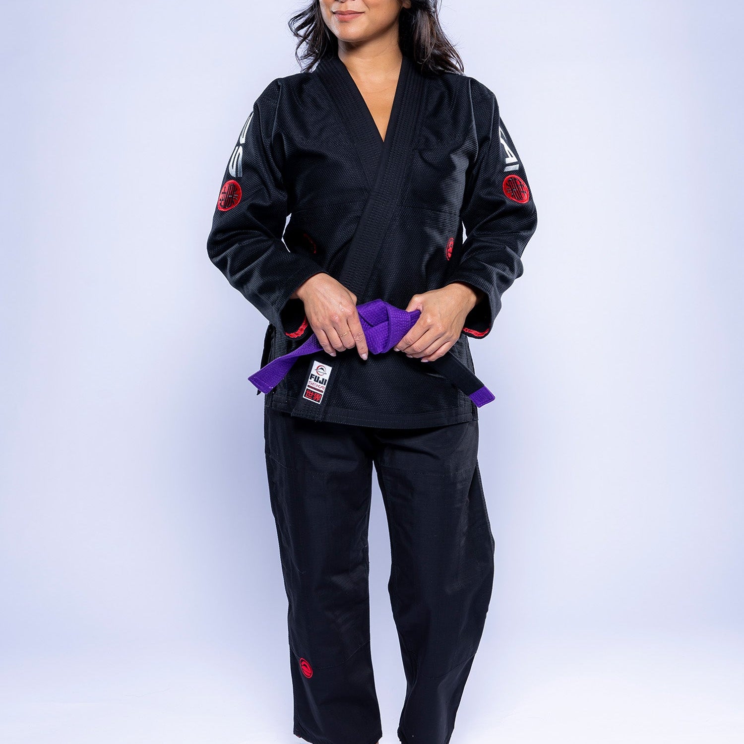 A woman in the Womens SEKAI 3.0 BJJ Gi Black Grey stands barefoot on a white background, holding a purple belt at her waist and looking to the side with a slight smile.