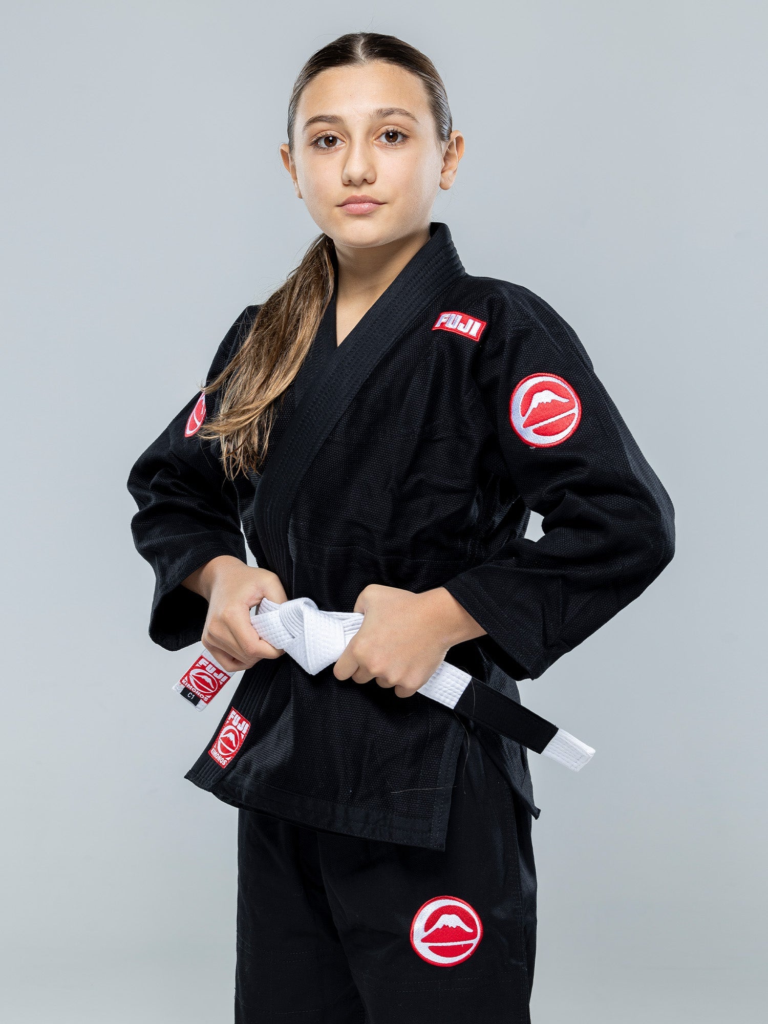 A young girl with long brown hair in a ponytail stands confidently wearing the Kids Competition BJJ Gi Black, featuring red and white patches, and holding a white belt against a plain gray background.
