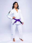 A woman with shoulder-length dark hair stands barefoot on a white background, smiling with her hands on her hips. She wears the Womens SEKAI 3.0 BJJ Gi White Teal and a purple belt tied at the waist.