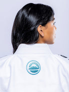 A woman with long dark hair is shown from behind wearing the Womens SEKAI 3.0 BJJ Gi White Teal, featuring a blue mountain and water emblem on the back of the collar as she faces to the right.