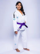 A woman stands barefoot on a white surface, confidently posing with one hand on her hip, wearing the Womens SEKAI 3.0 BJJ Gi White Teal with patches and a purple belt, smiling brightly.