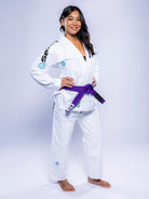A woman smiles barefoot on a white background, wearing the Womens SEKAI 3.0 BJJ Gi White Teal with a purple belt and her hands on her hips.