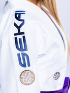A woman wears the Womens SEKAI 3.0 BJJ Gi White Navy with SEKAI embroidered in blue on the sleeve, round brown patches, a purple belt, and her blonde hair showing.