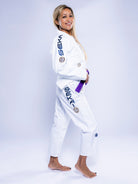 A woman smiles in profile, barefoot and wearing the Womens SEKAI 3.0 BJJ Gi White Navy with a purple belt. The gi features blue and black logos and patches on the arms and legs, set against a light background.